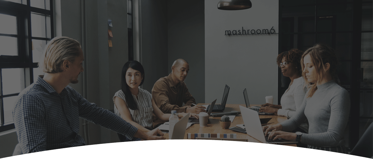 A group of five professionals engaged in a discussion at a meeting table with laptops.