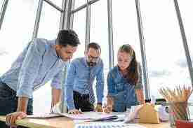 Three professionals reviewing project plans and documents at a table in an office setting.
