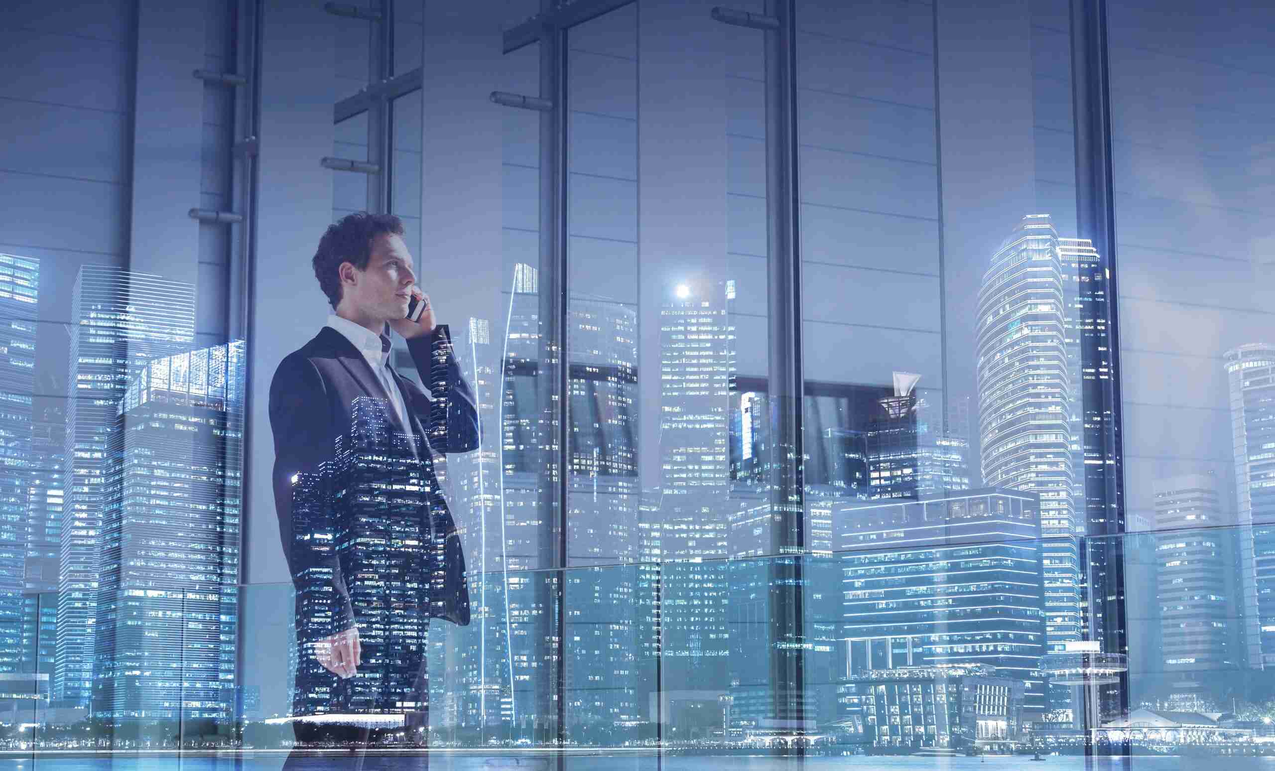 Businessman in a suit talking on a phone with a city skyline reflected in the glass behind him.