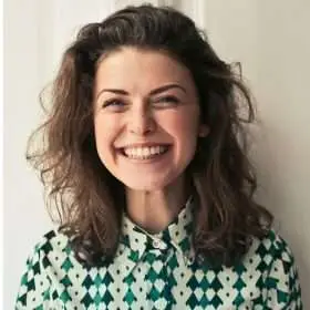 A woman with curly hair smiling while wearing a patterned shirt with green and white colors.