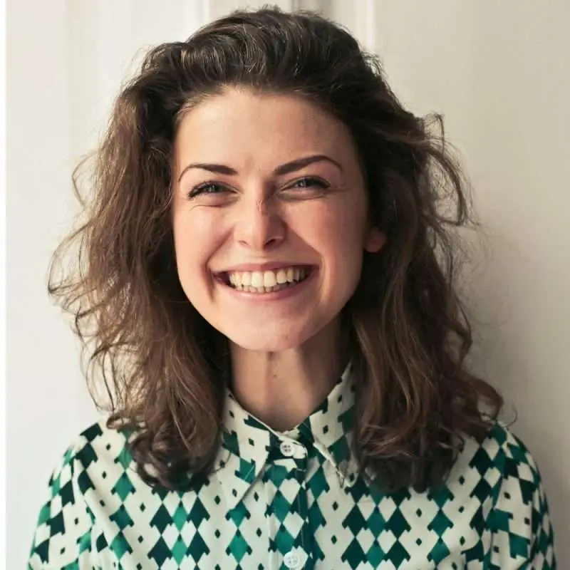 A woman with curly hair smiling while wearing a patterned shirt with green and white colors.